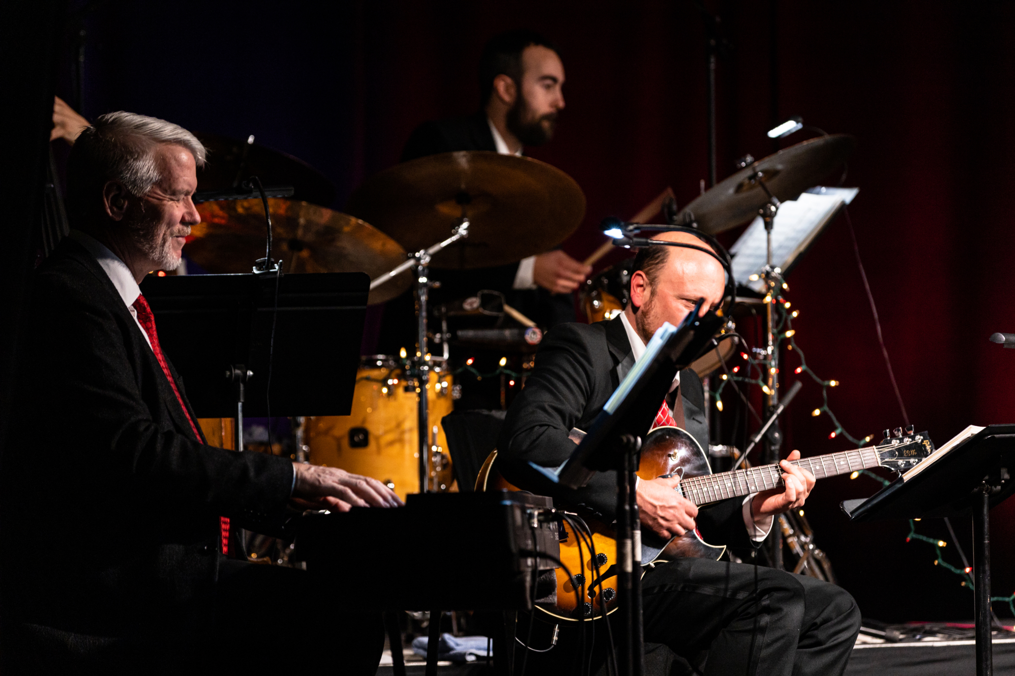 Profile view of three musicians playing seated on stage, a pianist in the foreground, a guitarist, and a drummer in the background.
