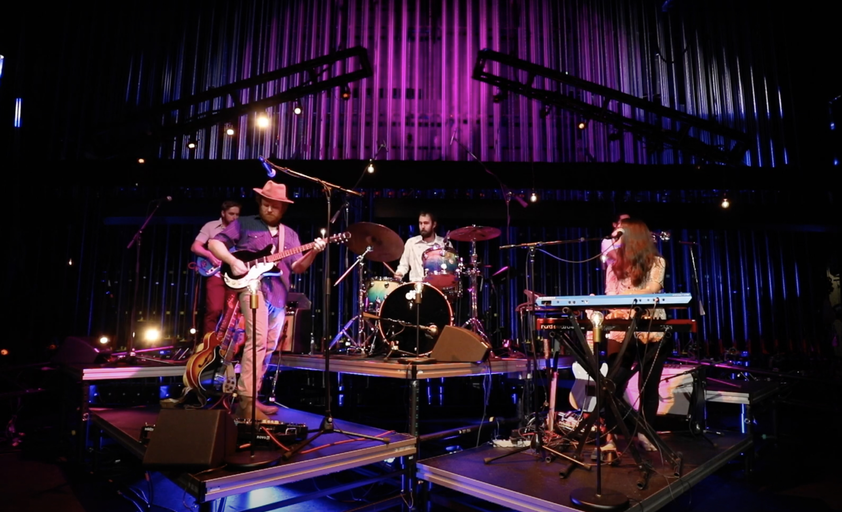 Band plays on a stage in colorful blue and purple lighting, four musicians playing various instruments on risers surrounding a drummer in the center.